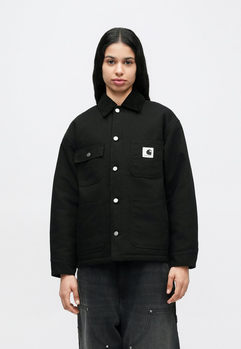 Persona joven con cabello negro largo que lleva una chaqueta negra abotonada con grandes bolsillos y pantalones oscuros y holgados sobre un fondo blanco liso.