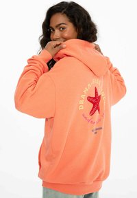 Korálová mikina s grafikou morskej hviezdice na chrbte. Obsahuje rebrované manžety a lem, mäkkú textúru a voľný strih pre pohodlie.