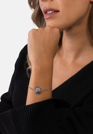 Frau trägt ein silbernes, sonnenförmiges Armband mit einem zentralen Edelstein am Handgelenk, gekleidet in ein schwarzes Langarmshirt, das Kinn auf die Hand gestützt.