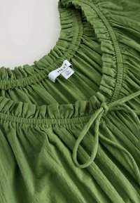 Vaatteet vihreä, jossa on rypistetty pääntie ja röyhelöyksityiskohtia. Valmistettu pehmeästä kankaasta, edessä solmittava vetskari ja "NEXT"-brändätty etiketti.