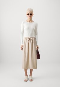 Haut blanc à manches longues avec une jupe midi à cordon beige, associé à des sandales à glisser de couleur claire et un petit sac à main bordeaux.