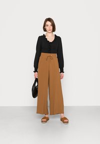 Blusa preta com decote franzido, calças largas marrons com cintura elástica e sandálias de plataforma marrons. Bolsa preta, fundo neutro.