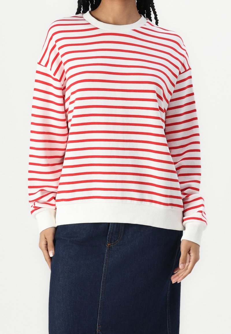 Personne portant une chemise blanche à manches longues avec des rayures rouges horizontales et une jupe en jean bleu foncé sur un fond blanc uni.