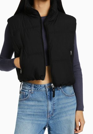 Frau trägt eine schwarze Steppweste über einem kurz geschnittenen, lila, gerippten Pullover und hoch taillierte hellblaue Jeans, mit einer Hand in der Westentasche.