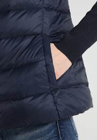 Námořnická péřová vesta s prošívaným designem, viditelná boční kapsa. Látka působí hladce s lehkým leskem, obsahuje žebrované manžetové detaily.