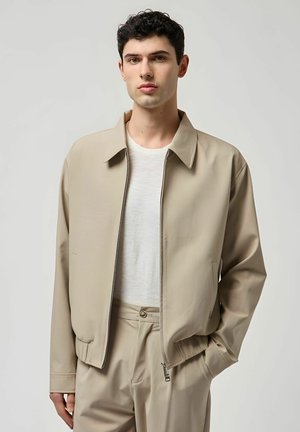 Giovane uomo che indossa una giacca beige con cerniera sopra una maglietta bianca e pantaloni beige abbinati, in piedi con una mano in tasca.