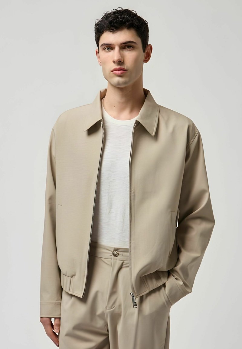 Giovane uomo che indossa una giacca beige con cerniera sopra una maglietta bianca e pantaloni beige abbinati, in piedi con una mano in tasca.