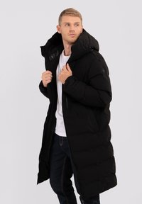 Giacca a vento nera con colletto alto, grande cappuccio e tasche con cerniera. Presenta una texture liscia e una vestibilità sagomata, indossata sopra una camicia bianca.