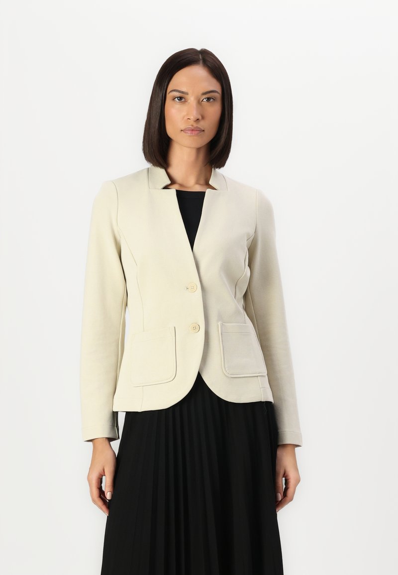 Blazer beige clair avec deux poches avant, un col simple et deux boutons. Le tissu a une texture subtile. Porté sur une robe noire.
