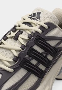 Baskets Adidas beige clair avec des supports de lacets noirs en forme de griffes et une tige en tissu maillé texturé.