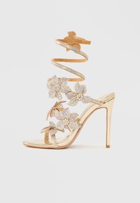 Kuldne kõrge kontsaga sandaal läikiva viimistlusega, lillemotiividega ja keeratud rihmaga. Kaunistatud kristallaktsentidega ning ühtlase tekstuuriga.