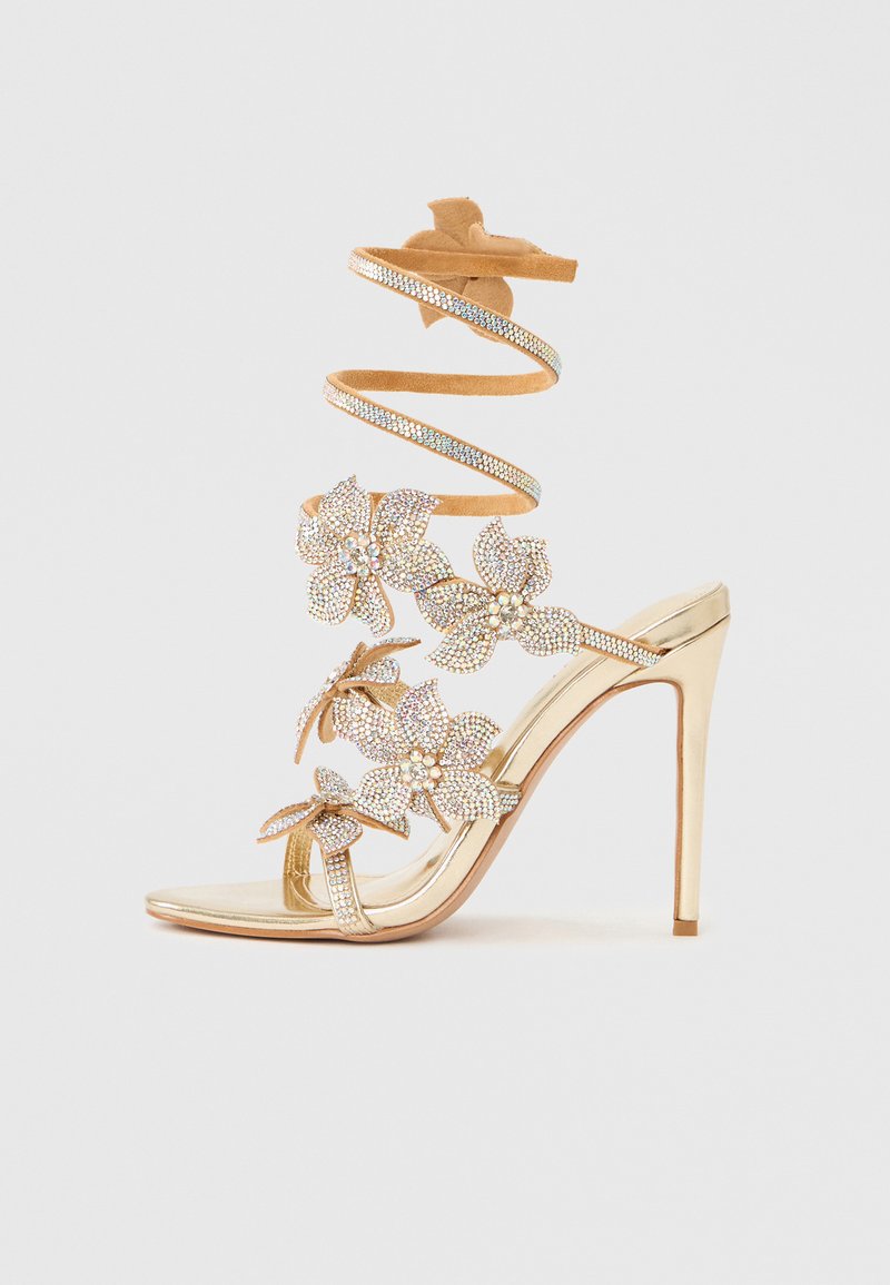 Kuldne kõrge kontsaga sandaal läikiva viimistlusega, lillemotiividega ja keeratud rihmaga. Kaunistatud kristallaktsentidega ning ühtlase tekstuuriga.