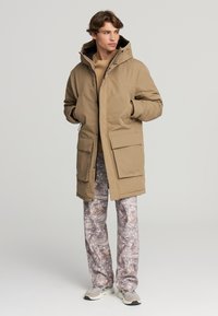 Giacca parka beige con cappuccio, dotata di ampie tasche a toppa e chiusura con zip, abbinata a pantaloni grigi a fantasia e sneakers grigie.
