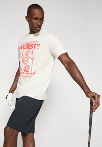 Cream-colored cotton t-shirt with red graphic text and cartoon hotdog design. Paired with dark shorts and wearing a white golf glove.