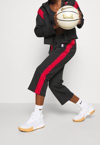 Sort træningsjakke med røde striber, matchende sorte cropped bukser med røde striber, hvide sneakers med et struktureret design, holder en basketball.