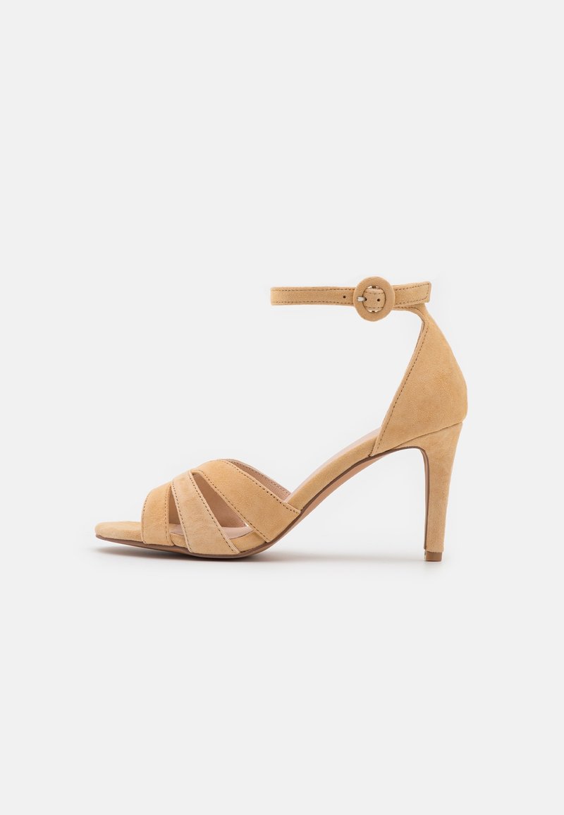Sandale beige en daim à talon haut avec bout ouvert, trois brides à l'avant, bride à la cheville et boucle ronde sur fond blanc uni.