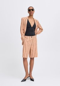 Costume rayé pêche avec blazer ajusté et shorts, associé à un maillot de bain noir et des escarpins noirs à bout pointu, des lunettes de soleil accentuent le look.