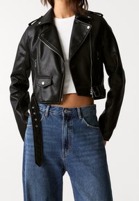 Chaqueta de motociclista corta de cuero negro con herrajes plateados, cremallera frontal, bolsillo lateral y detalle de correa ajustable, combinada con jeans azul de cintura alta.