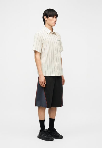 Νεαρός άνδρας που στέκεται, φοράει λευκό ριγέ πουκάμισο με κοντά μανίκια και κουμπιά, μαύρα και γκρι σορτς με κόκκινες λεπτομέρειες, μαύρες κάλτσες και μαύρα αθλητικά παπούτσια.