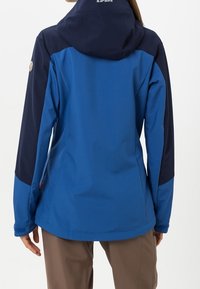 Femme portant une veste d'extérieur à capuche bleue et marine et un pantalon marron, vue de dos sur un fond blanc.