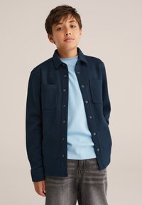 Mornarsko modra srajca-jakna s gumbi in dvema sprednjima žepoma, teksturirana tkanina in ovratnik, nosi se čez svetlo modro majico in sive kavbojke.