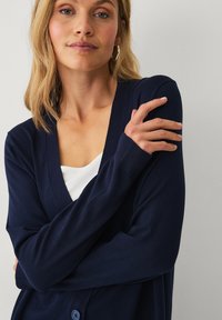Donkerblauwe cardigan met een v-hals ontwerp, geribbelde boorden en een knoopdetail. Draagt over een witte top, tegen een lichte achtergrond.