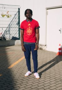 Κόκκινο κοντομάνικο μπλουζάκι με το σήμα της Ισπανίας, συνδυασμένο με ναυτικές παντελόνες και λευκά αθλητικά παπούτσια με κόκκινες λεπτομέρειες. Υπαίθριος χώρος με επίπεδη επιφάνεια σε πλέγμα.