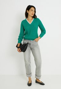 Suéter verde de punto con escote en V y borde festoneado, combinado con jeans grises con puños y bolso bandolera negro. Incluye zapatos planos negros con lunares blancos.