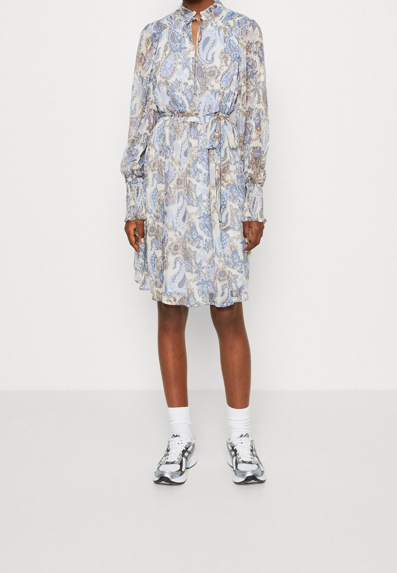 Robe à motifs cachemire bleu clair avec des manches longues, des poignets froncés, un col haut et une ceinture à nouer. Portée avec des chaussettes blanches et des baskets argentées.