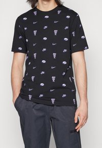 Černé tričko s krátkým rukávem s opakujícími se fialovými logy Nike, swooshy a textem "Just Do It", nošené s tmavě šedými kalhotami.