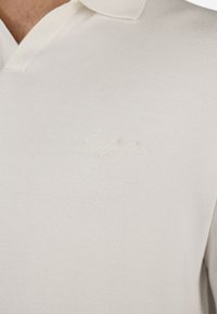 White cotton polo shirt with a collared neck, featuring a subtle embroidered logo on the chest. Smooth texture and classic design.