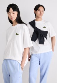 Dos personas llevan camisetas blancas con el logo del cocodrilo verde de Lacoste. La mujer combina su camiseta con pantalones de chándal azul claro; el hombre tiene un suéter oscuro atado a la cintura.