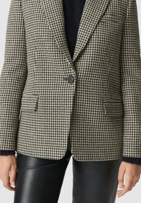 Blazer a motivo pied de poule beige e nero, con un bottone singolo, revers a taglio e due tasche frontali. Abbinato a pantaloni in pelle nera.