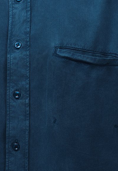 Κοντινό πλάνο υφάσματος μπλε πουκάμισου με κουμπιά, που δείχνει τσέπη welt στο αριστερό στήθος και τέσσερα μπλε κουμπιά στην περιοχή του πασαλείμματος.