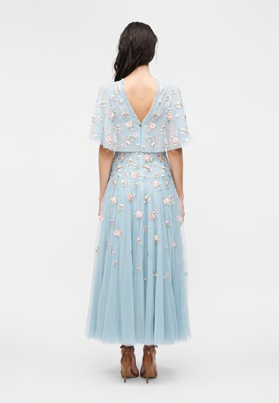 Kvinde med langt mørkt hår iført en lyseblå, blomsterbroderet kjole med transparente korte ærmer og åben ryg, kombineret med brune hæle.