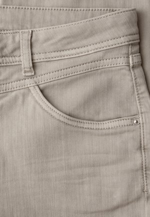 Nærbillede af beige denimjeans' forlomme med synlig bæltestrop, syninger og lille metalnitte.