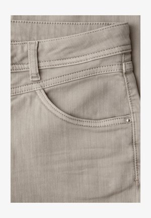 Nærbillede af beige denimjeans' forlomme med synlig bæltestrop, syninger og lille metalnitte.