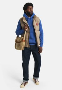 Beige polstret vest over en blå hættetrøje, mørkeblå jeans og beige sneakers. Bærer en taske i tan og brun over skulderen.