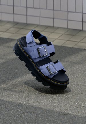 Vastag fekete talpú szandál, levendulaszínű dupla csatos pántokkal, texturált járdán pihen, csempézett fal háttérrel.