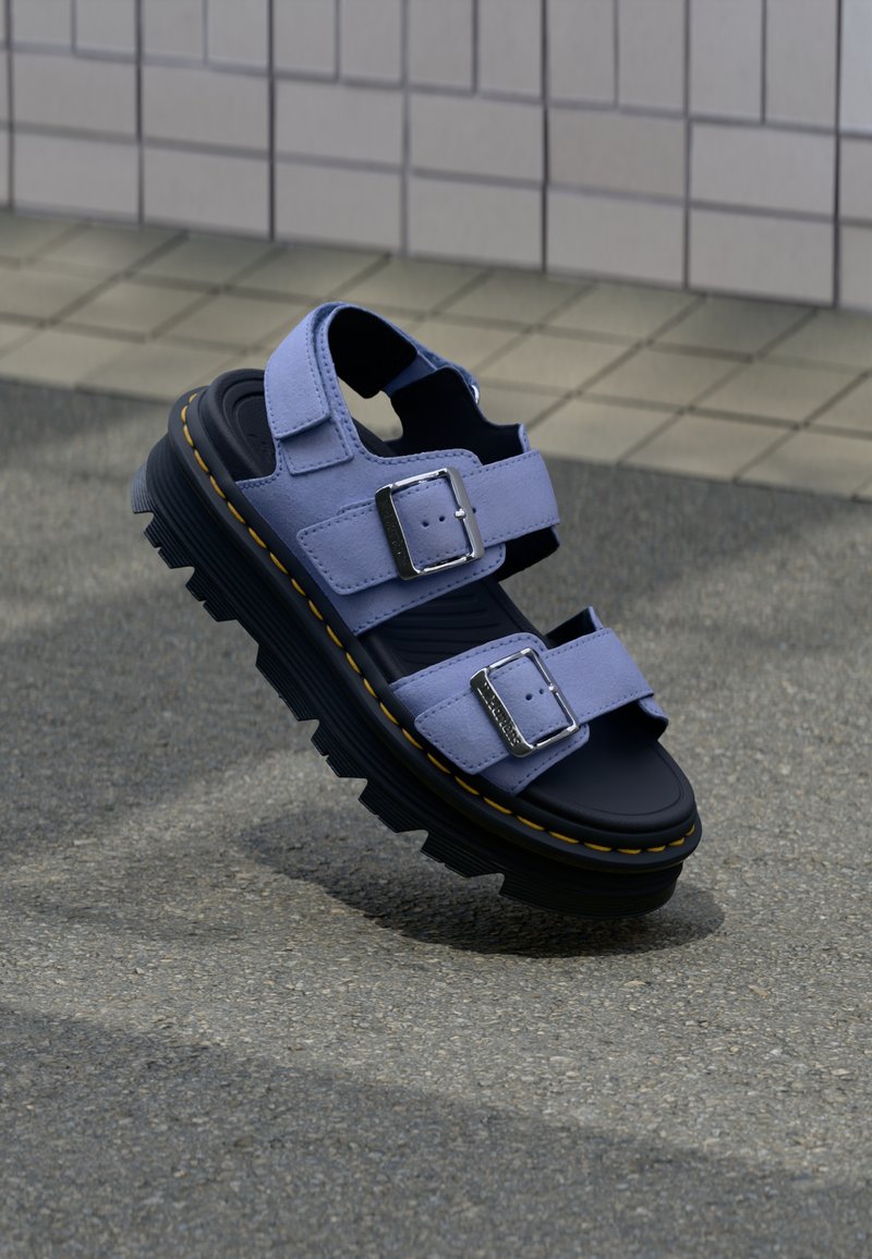 Πέδιλο με παχιά μαύρη σόλα και διπλούς ιμάντες με αγκράφες σε χρώμα λεβάντας, ακουμπισμένο σε υφή άσφαλτου κοντά σε φόντο από πλακιδωτούς τοίχους.