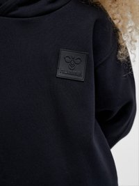 Zbliżenie na czarną bluzę z kapturem z wytłoczonym naszywką z logo hummel na piersi, noszoną przez osobę o kręconych jasnych włosach.