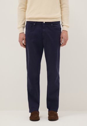 Mann trägt dunkelblaue Hose, beigen Pullover und braune Wildleder-Loafer, steht vor einem einfarbigen hellen Hintergrund.