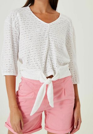 Femme portant un chemisier blanc à nouer devant en broderie anglaise avec des manches trois-quarts et un short rose à ourlet roulé, debout devant un fond neutre.