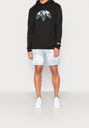 Osoba w czarnej bluzie z kapturem z jasnoniebieskim geometrycznym logo, jasnoszarych szortach tie-dye i białych tenisówkach, stojąca na gładkiej podłodze.