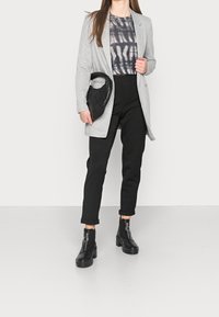 Šedý blazer přes černé top s tie-dye vzorem, v kombinaci s černými zúženými kalhotami a robustními černými zipovými botami; drží černou prošívanou kabelku.