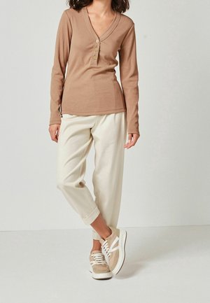 Persoon draagt een lichtbruine lange mouwen V-hals top met knopen, crème losse broek en beige witte sneakers, staand op een witte vloer.