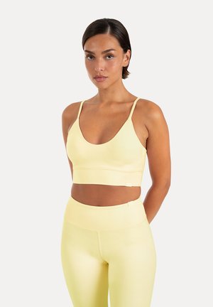 Femme aux cheveux courts et foncés portant un soutien-gorge de sport jaune pâle et un legging taille haute assorti, debout devant un fond clair uni.