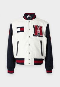 Giacca varsity bianca con maniche blu navy, caratterizzata da polsini a coste rossi e blu navy, toppe con logo e bottoni a pressione. Texture liscia in pelle e maglia.