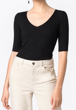 Femme portant un haut noir côtelé à col en V avec des manches mi-longues et un pantalon taille haute écru, debout devant un fond uni.