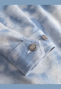 Light blue denim fabric with a tie-dye effect. Features a close-up of a cuff with two decorative metallic buttons.